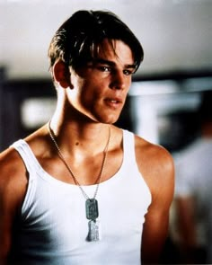 a young man wearing a white tank top with a tassel on his neck and looking at the camera