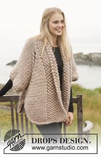 a woman wearing a knitted cardigan standing next to a wooden fence with grass and water in the background