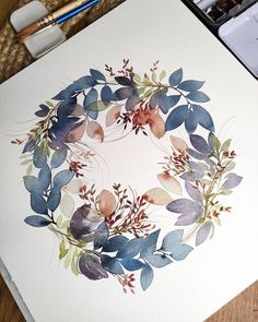 a watercolor painting of a wreath made with leaves and berries on white paper next to paintbrushes