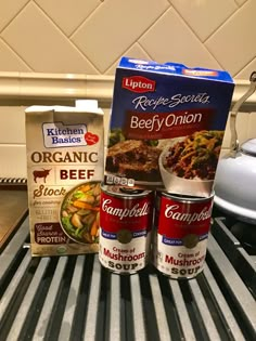 three cans of soup sitting on top of a grill next to an open box of canned food