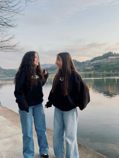 two young women standing next to each other near the water with their hands in their pockets