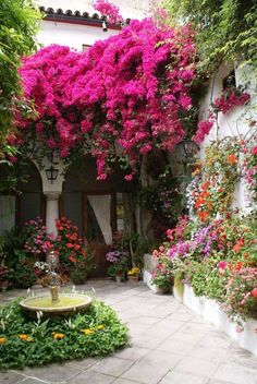 an outdoor garden with lots of flowers growing on the walls and around it is a fountain surrounded by greenery