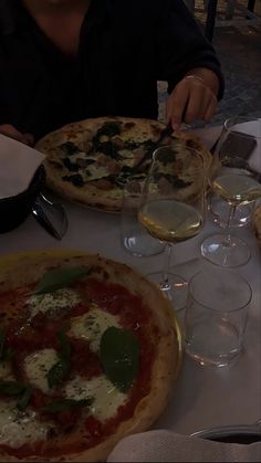 two pizzas and wine glasses on a table with people sitting at the table eating