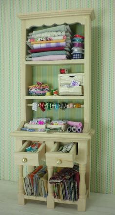 a dollhouse shelf with many items on it and some folded fabric in the bottom drawer