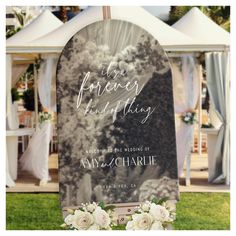 a wedding ceremony sign with flowers on the grass in front of a tented area