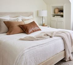 a bed with white linens and brown pillows in a small room next to a window