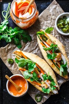 two fish tacos are sitting on wax paper with dipping sauces and vegetables in the background