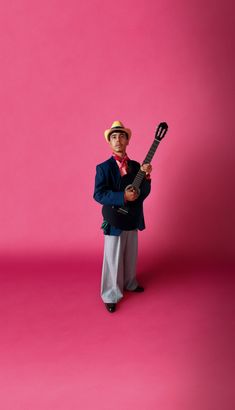 a man in a suit and hat holding a guitar against a pink background with his arms crossed