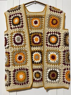 a crocheted vest is hanging on a white wooden wall and it has an orange, red, yellow, and brown flower motif