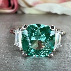 "The ring pictured is lab created green paraiba with moissanite accents #6778 This ring can be made with many different stone colors Message us for pricing on different color/type stones -Approximate total carat weight:  2.80ctw diamond equivalent  -Center Stone Size: 8x8mm - approx. 2.40ct diamond equivalent -Gem Type:  lab created Green Paraiba -Center Stone Shape:  Cushion cut  -Center Stone Color: green (mint green) -Center Stone Clarity: VS2 -Moh's Scale: 7.5 - 8 hardness -Side Stone Size: Paraiba Tourmaline Ring, Jewellery Board, Ring Three Stone, Green Tourmaline Ring, Gold Cushions, Paraiba Tourmaline, White Gold Set, Green Mint, Mothersday Gifts