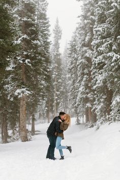 couple is sharing a kiss in the middle of a snow covered pine forest, woman is popping her foot Winter Engagement Photos Snow, Utah Winter, Cute Engagement Photos