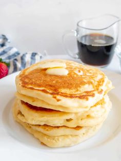 a stack of pancakes on a white plate with syrup and strawberries in the background