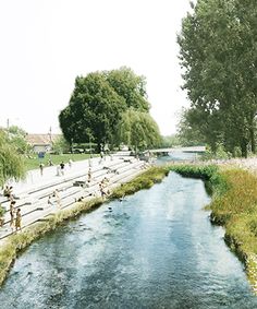 a river running through a lush green park next to tall grass covered trees and people standing on the bank