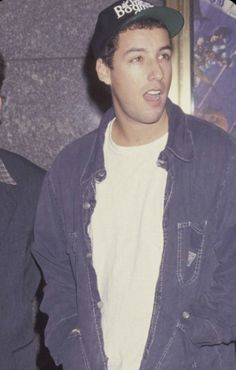 a man wearing a hat standing next to another man in a blue jacket and white t - shirt