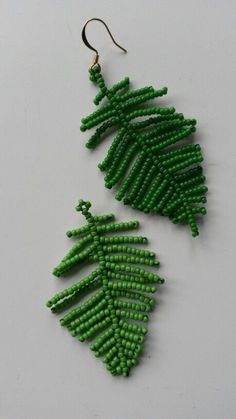 two green beaded earrings hanging from hooks on a white surface, one has a fern leaf design