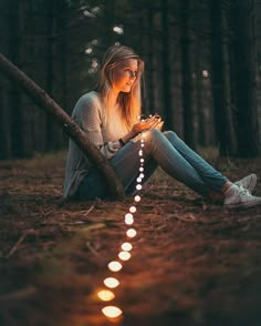 a woman sitting on the ground in the woods looking at her cell phone while holding a string of lights