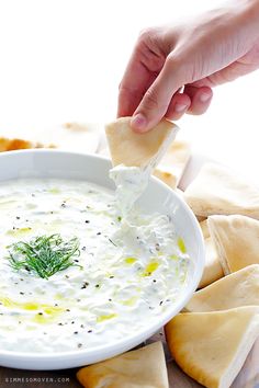 a person dipping dip into a white bowl with pita bread on the side and green garnish