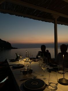 two people sitting at a table with wine glasses and plates in front of the ocean