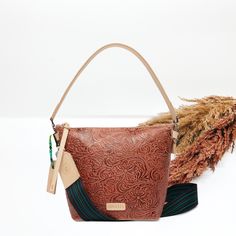 A wedge tooled bag sits in the middle of the picture in a brown tooled pattern. Background is solid white with pompas grass to the right of the purse. Western Style Brown Hand Tooled Shoulder Bag, Consuela Crossbody Bags, Southwestern Style Bag With Adjustable Strap, Western Leather Bags With Concho Details, Brown Hand Tooled Crossbody Shoulder Bag, Leather Luggage Tags, Giddy Up Glamour, By Your Side, Leather Luggage