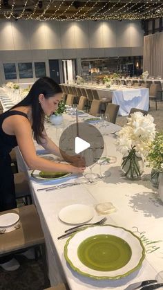 a woman is setting the table for a formal dinner with white flowers and greenery