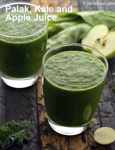 two glasses filled with green smoothie on top of a wooden table next to sliced apples