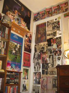 a wall covered in posters and pictures next to a dresser with a lamp on it