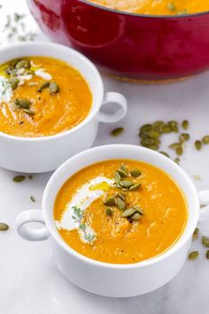 two white bowls filled with carrot soup and topped with pumpkin seeds