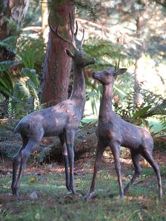 two statues of deer standing next to each other in the grass near trees and bushes