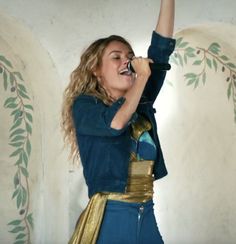 a woman dressed in blue and gold singing into a microphone with her hand raised up