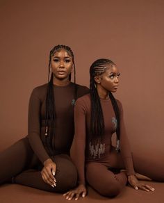 two women sitting on the ground with long braids in their hair and wearing brown bodysuits