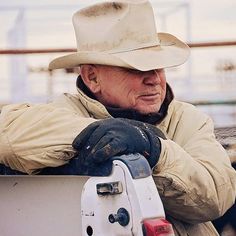 an old man wearing a cowboy hat and gloves leaning on the back of a truck