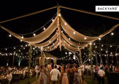 a group of people standing under a tent covered in string lights and draped with white fabric