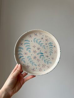 a hand holding a white plate with blue and pink flowers painted on the side, in front of a gray wall