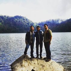 three men standing on a rock in the middle of a body of water with mountains in the background