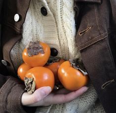 a person holding several orange tomatoes in their hands and wearing a brown jacket with buttons