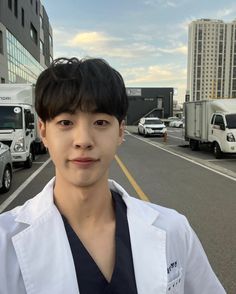 a male in a white lab coat standing on the side of a road with trucks behind him