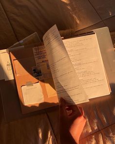 someone is reading a book on their bed with the sun shining through it and there are papers in front of them