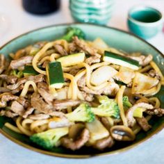 a green bowl filled with noodles and vegetables on top of a white table next to cups