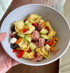 a person holding a white bowl filled with pasta and veggies on top of a wooden table