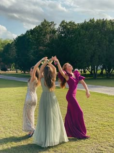 three women in long dresses are holding their hands up to the sky while standing on grass