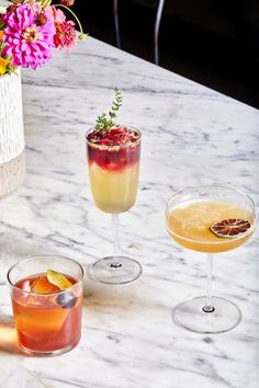 three cocktails on a marble table with flowers in vases and glasses filled with drinks