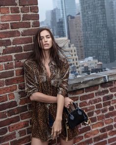 a woman leaning against a brick wall with her hand in her pocket and holding a purse