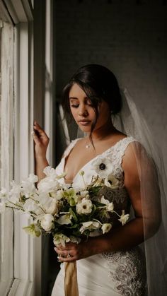 a woman in a wedding dress standing by a window holding a bouquet of white flowers