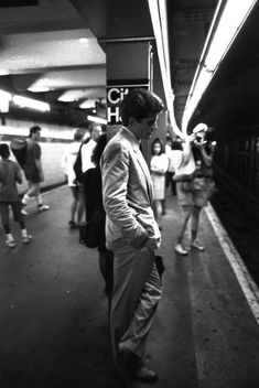 a man standing on a subway platform with his hands in his pockets as people walk by