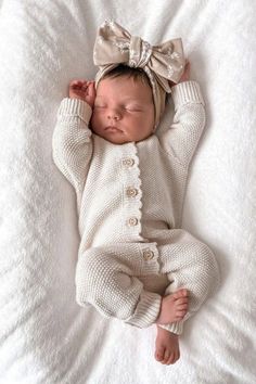 a baby is laying down wearing a white outfit and a bow on its head, with her hands behind her head