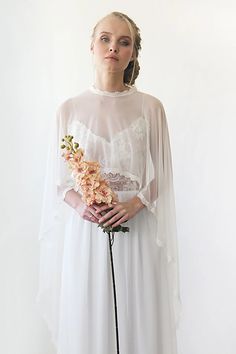 a woman wearing a white dress and holding a flower in her hand, standing against a wall