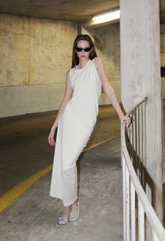 a woman leaning against a rail in a parking garage wearing sunglasses and a white dress