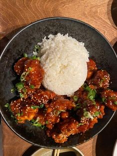 a black plate topped with meat and rice on top of a wooden table next to chopsticks