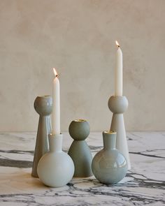 three white candles sitting next to each other on top of a marble table with two vases and one candle holder