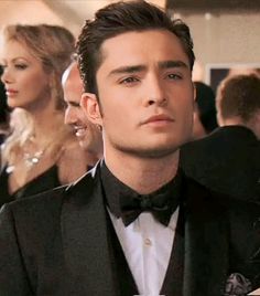 a man in a tuxedo is looking at the camera with other people behind him